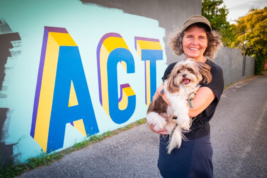 Christina Pollard holding her dog in front of an Act Belong Commit graffiti mural.