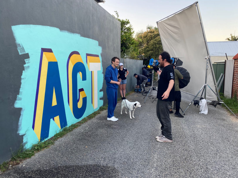 A behind the scenes photoshoot image with a man standing in front of the word 'Act' painted on a wall