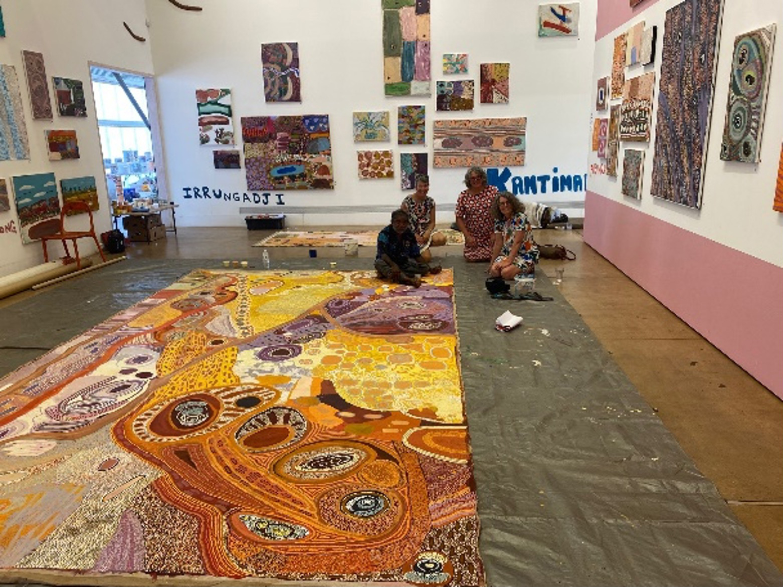 Four people sitting on the floor of an art gallery behind a large aboriginal painting